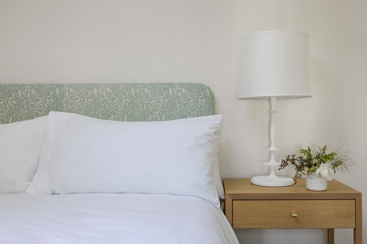A neatly made bed with white linens and pillows, a bedside table with a white lamp and a small vase with flowers, and a patterned headboard.