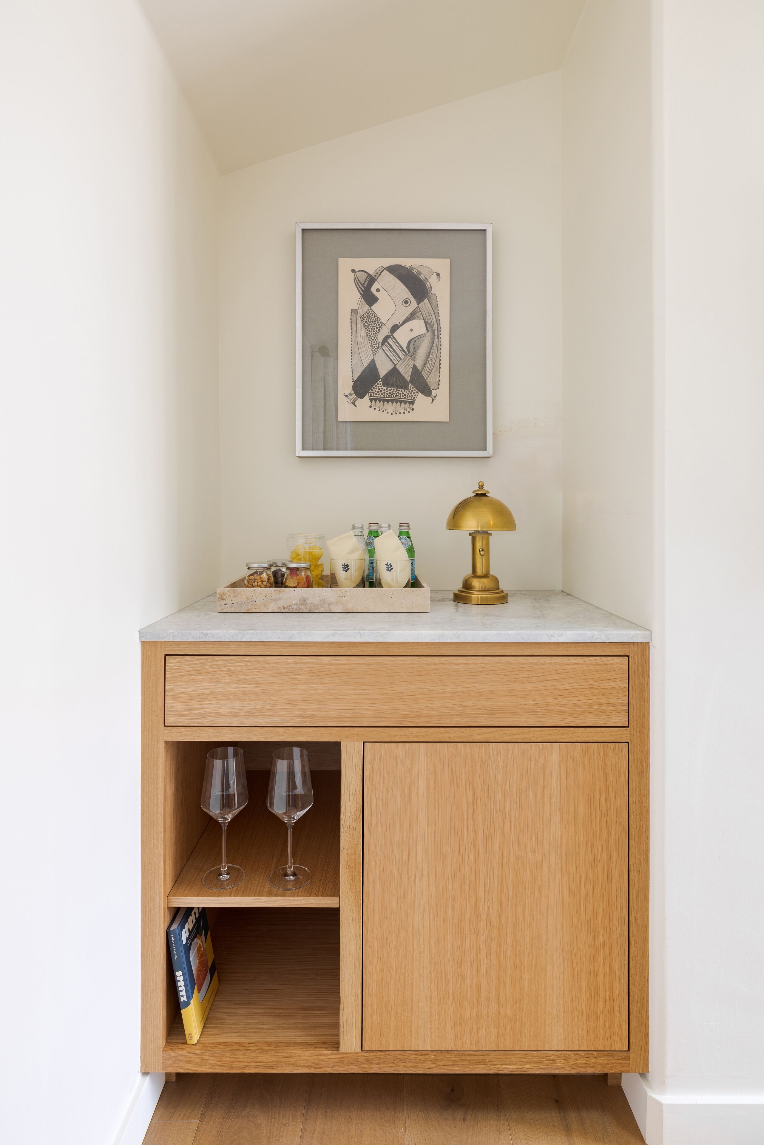 A wooden cabinet with a marble countertop, a framed artwork, a gold lamp, and two wine glasses.