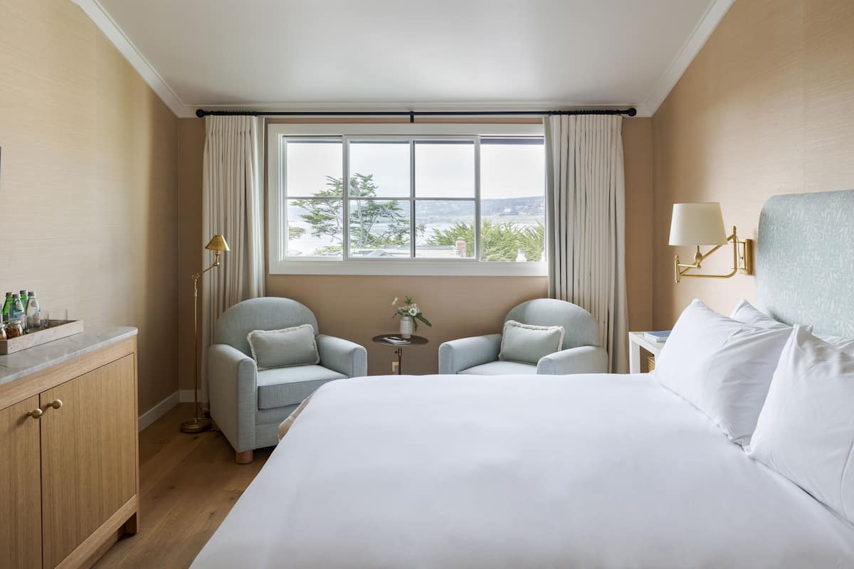A hotel room with a large bed, two armchairs, a small table with a vase, and a window with curtains.