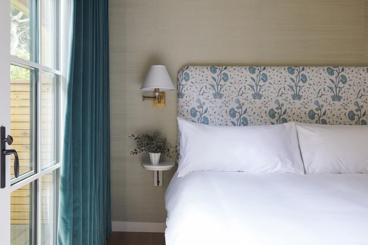 A neatly made bed with white linens, a wall-mounted lamp, and a small bedside table with a plant.