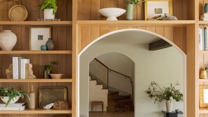 Interior de madera con un arco central que da a una escalera. Estanterías a ambos lados exhiben diversos objetos decorativos y libros.