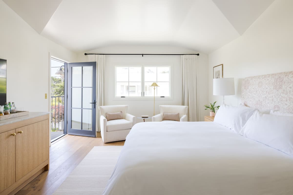 A hotel room with a large bed, two armchairs, a small table, a desk with a lamp, and a view through a glass door.
