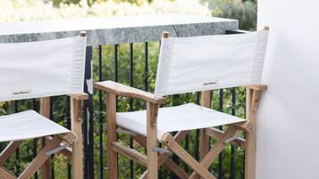 Two wooden chairs with canvas seats and backs, positioned on a wooden deck with a railing, overlooking a garden with trees and a building in the background.
