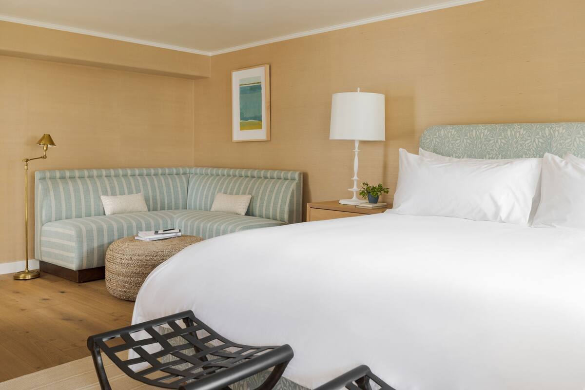 A hotel room with a large bed, a striped sofa, a small round ottoman, a black chair, a floor lamp, and a nightstand with a lamp and a plant.