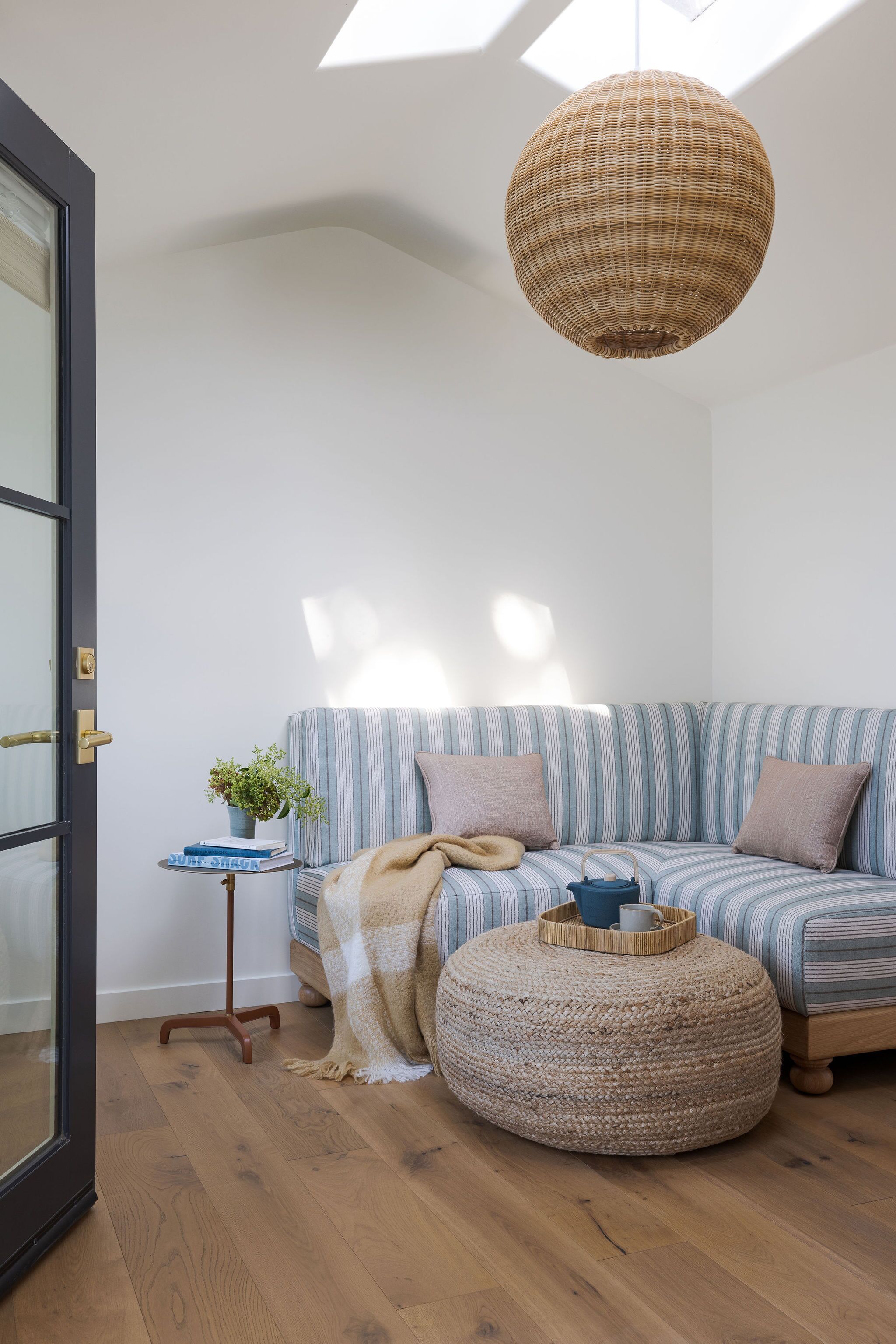 Ein modernes Wohnzimmer mit einem gestreiften Sofa, einem gewebten Pouf, einem kleinen Beistelltisch mit Pflanze und einer großen, gewebten Pendelleuchte.