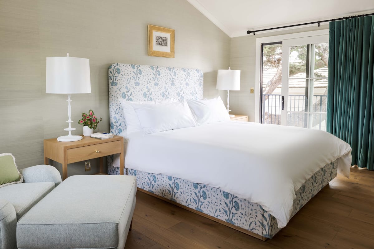 A neatly arranged bedroom with a large bed, two bedside tables with lamps, a small bedside table with a plant, and a view of the outdoors through a sliding door.