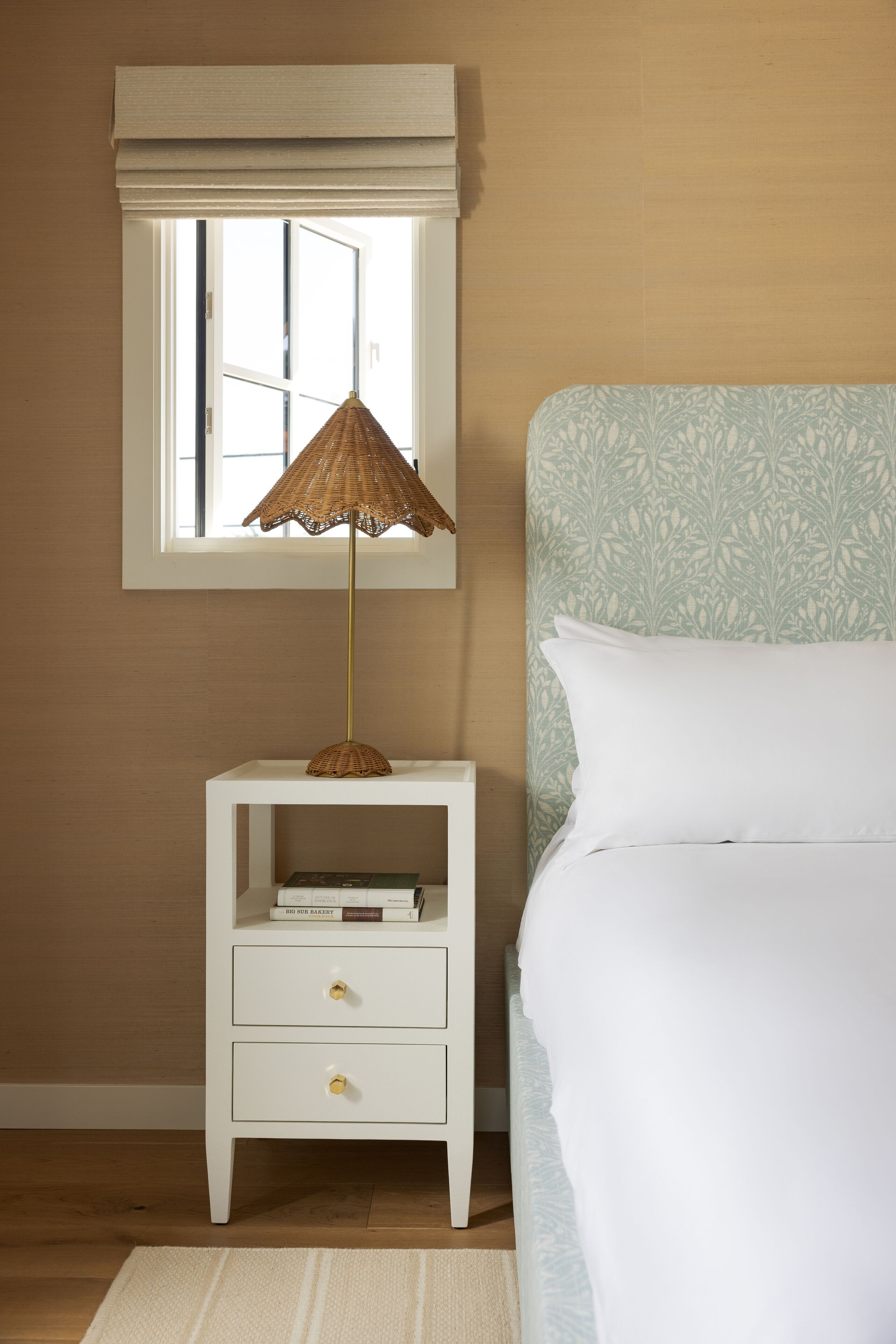 A bedroom with a white nightstand, a wicker floor lamp, a window with blinds, a patterned wall, and a bed with white bedding.