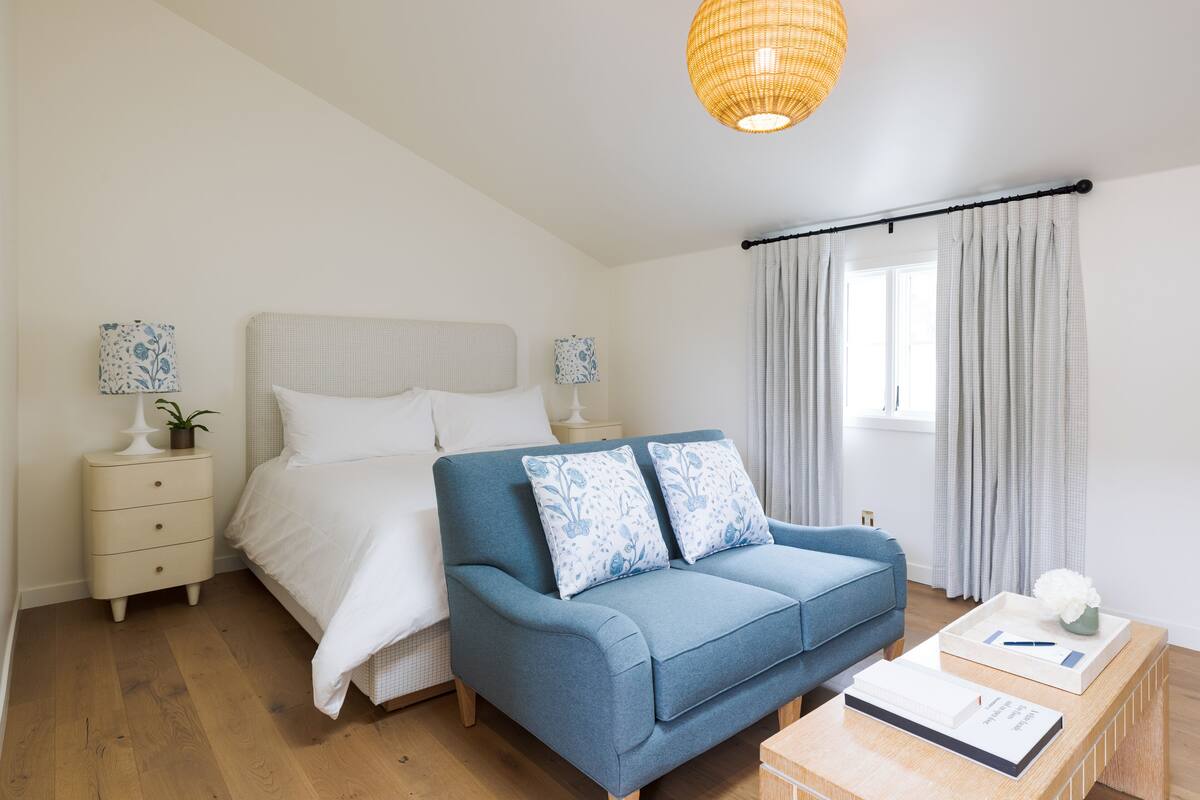 A bedroom with a bed, a blue sofa, a bedside table with a lamp, and a window with curtains.