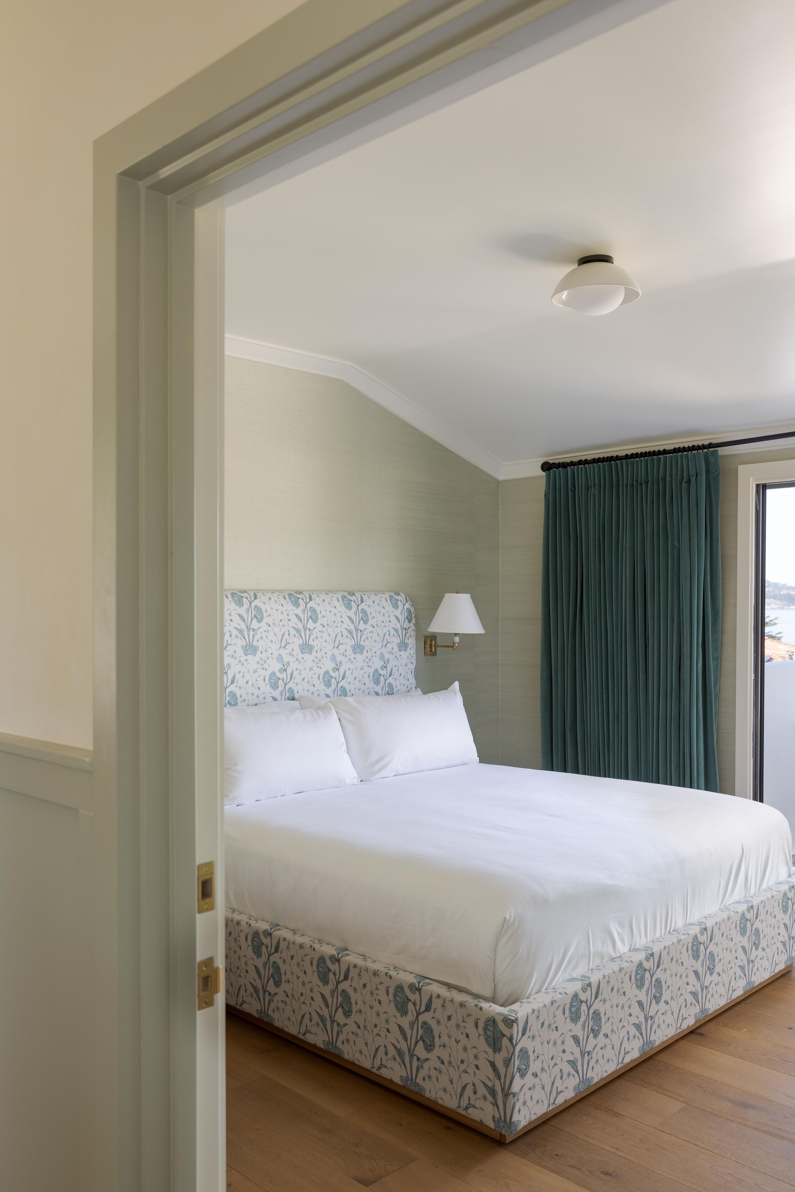 A bedroom with a bed, a nightstand, a lamp, and a view of the outside.