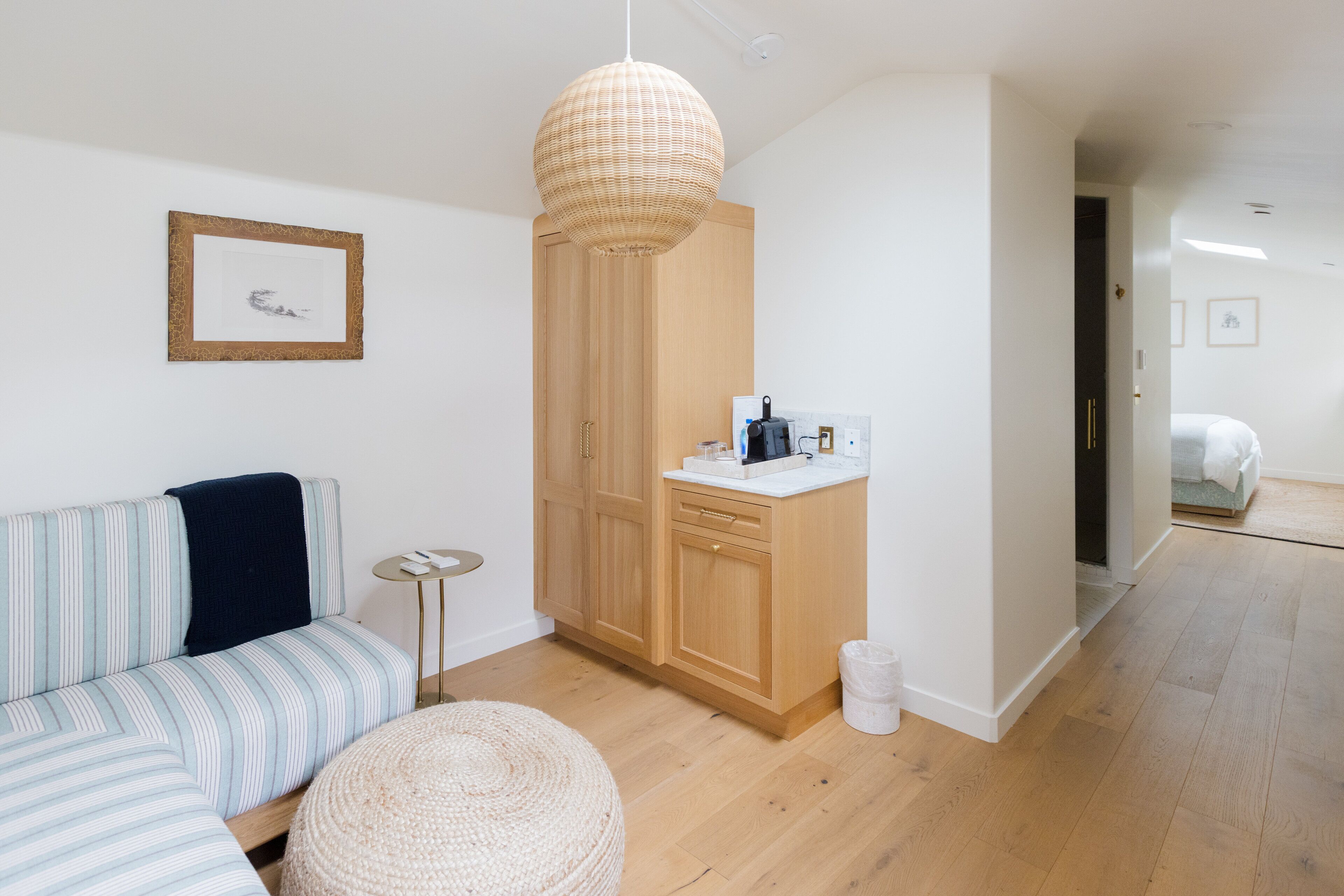 Una habitación moderna y bien iluminada, con un sofá a rayas, un otoman tejido redondo, un armario de madera y una pequeña cocina americana.