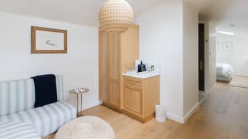 Una habitación moderna y bien iluminada, con un sofá a rayas, un otoman tejido redondo, un armario de madera y una pequeña cocina americana.