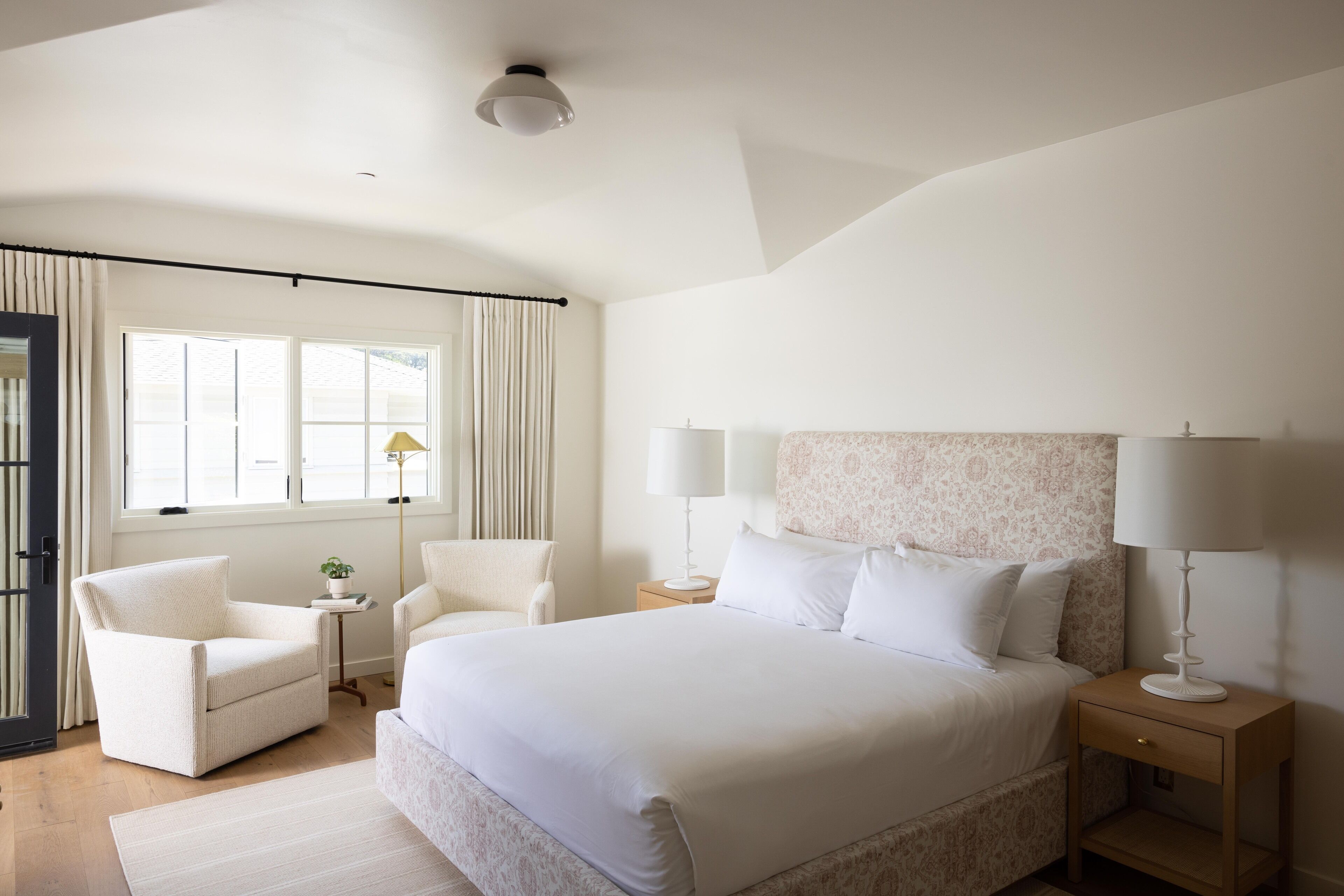 A spacious bedroom with a large bed, two bedside tables, a chair, and a window with curtains.