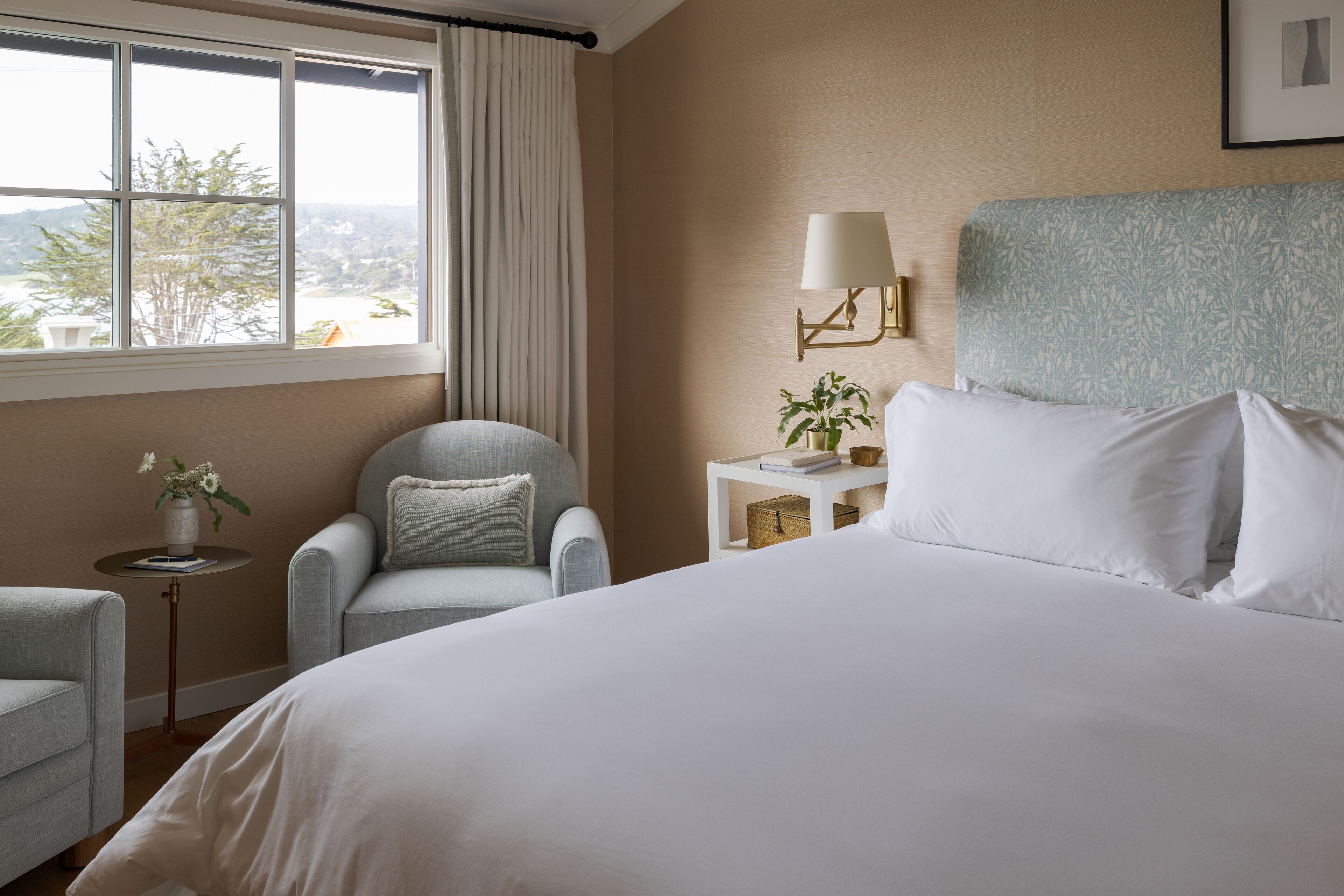 A hotel room with a large bed, a chair, a bedside table with a lamp, and a view through a window.