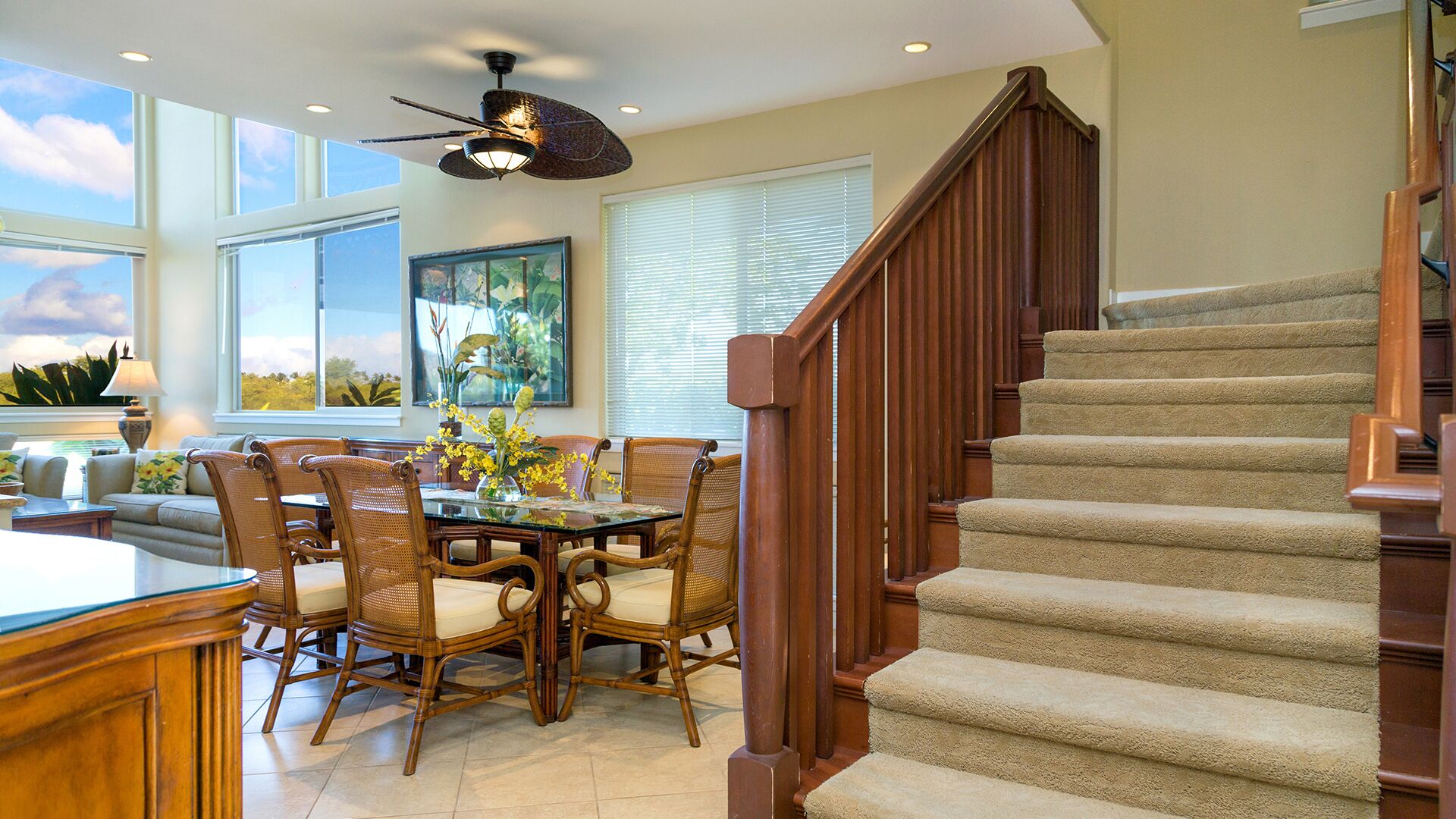 A dining area with a glass-top table, wicker chairs, and a view of the outdoors through large windows.