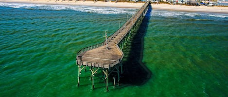 An aerial view of a pier extending into clear green waters, with a beach and buildings in the background.