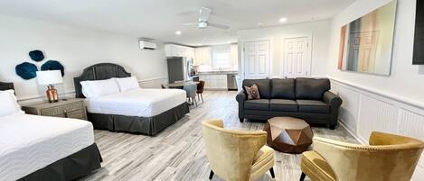A hotel room with a large bed, a sofa, two armchairs, a coffee table, and a kitchenette in the background.