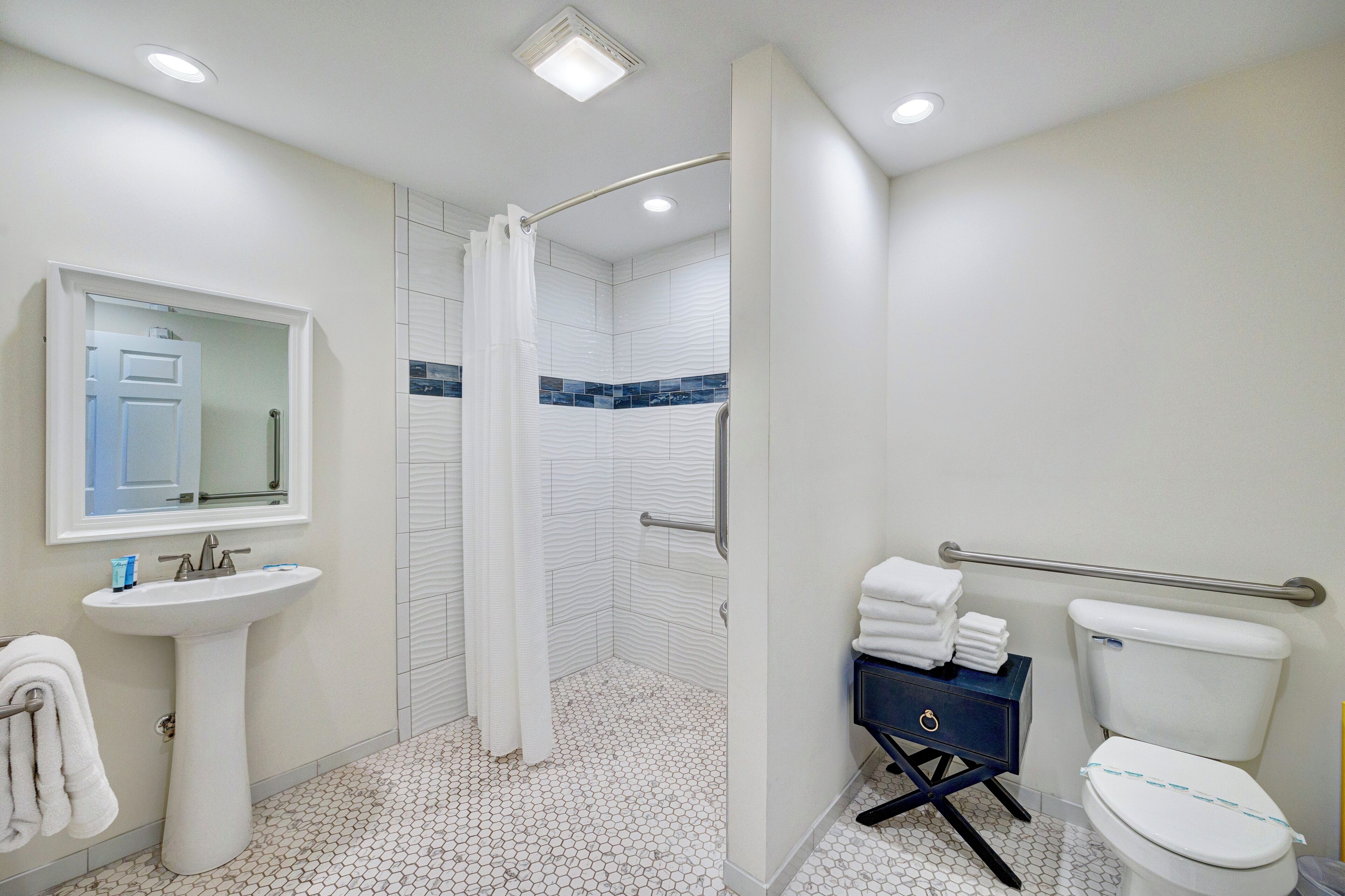 Un baño con lavamanos blanco, un espejo, ducha con puerta curva, inodoro con barra de apoyo y un armario azul.