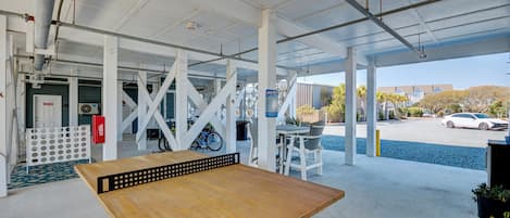 A covered outdoor seating area with a wooden table, white columns, and a view of a parking lot and buildings in the background.