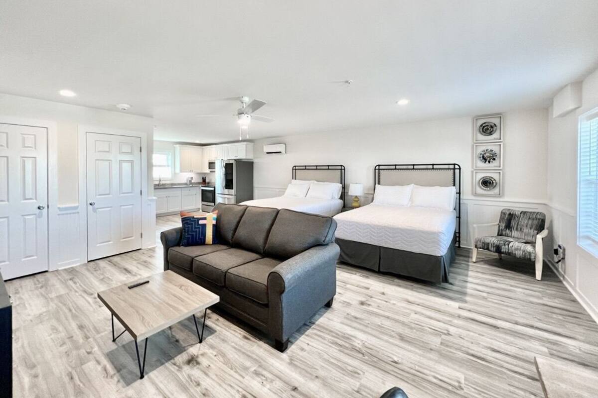 A modern hotel room with a sofa, two beds, a dining table, and a kitchenette.