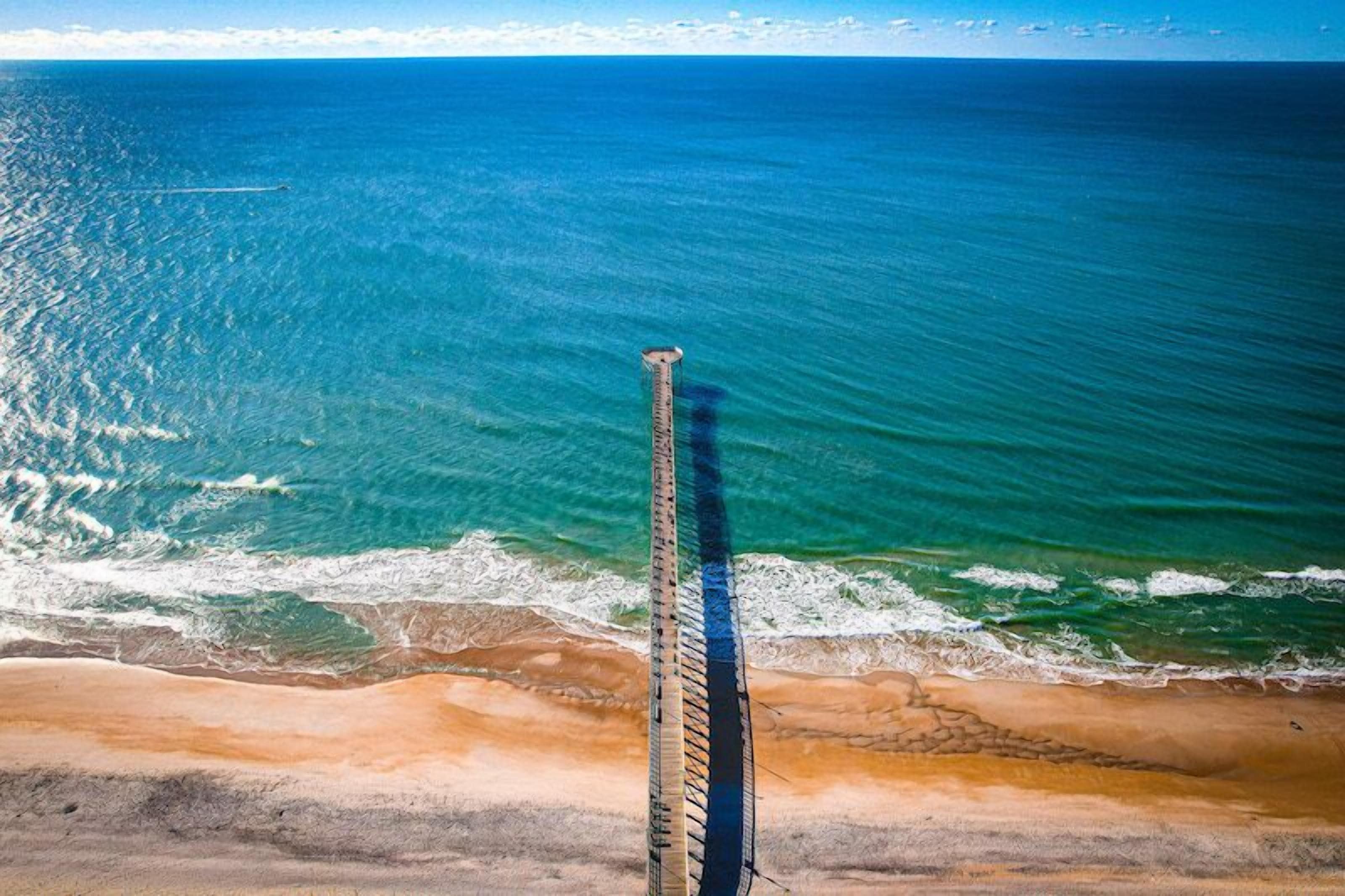 Un paysage côtier avec une longue jetée étroite qui s’avance dans l’océan, entourée d’une plage de sable et d’une eau bleue et limpide.