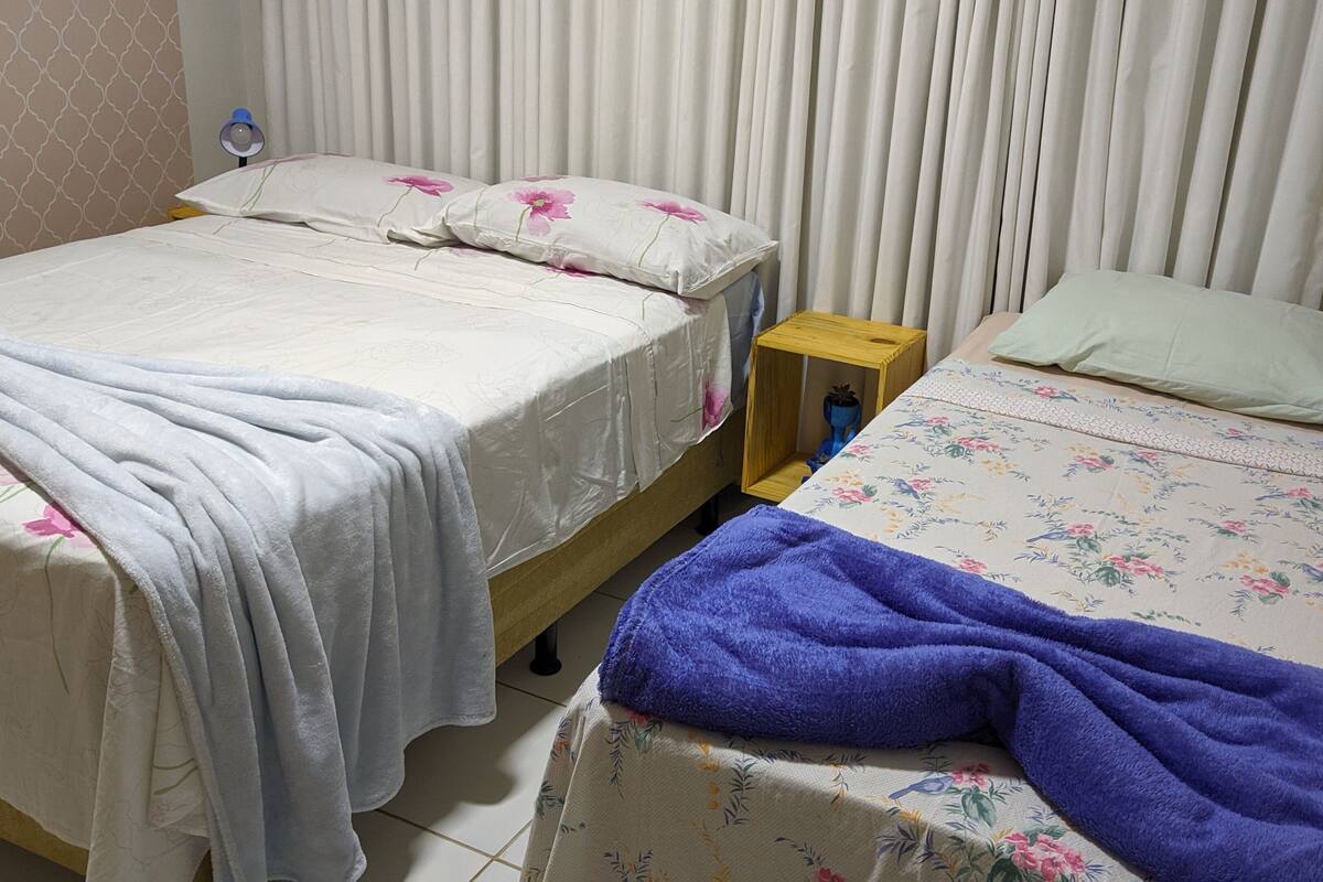 A bedroom with two beds, one with a floral bedspread and the other with a plain one. There is a wooden nightstand with a lamp between the beds.