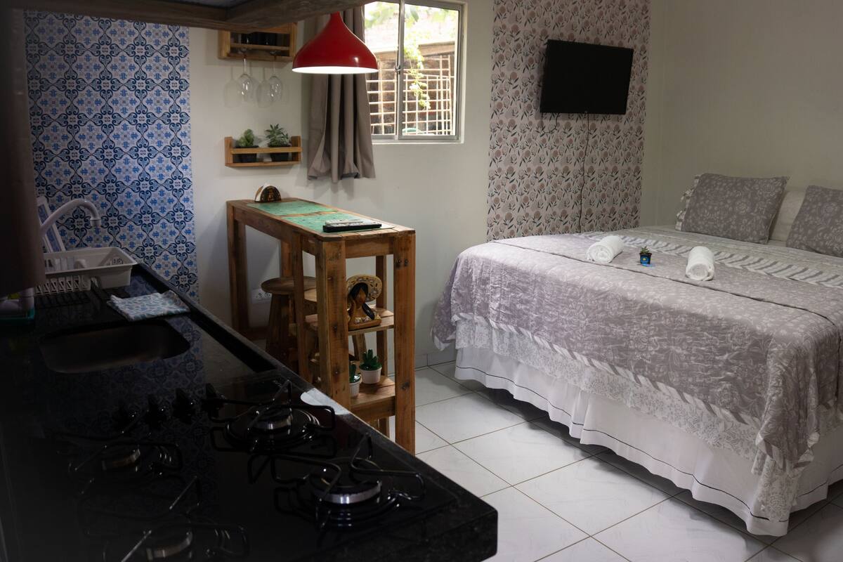 A bedroom with a bed, pillows, and a bedside table. There is a window with curtains and a TV mounted on the wall.