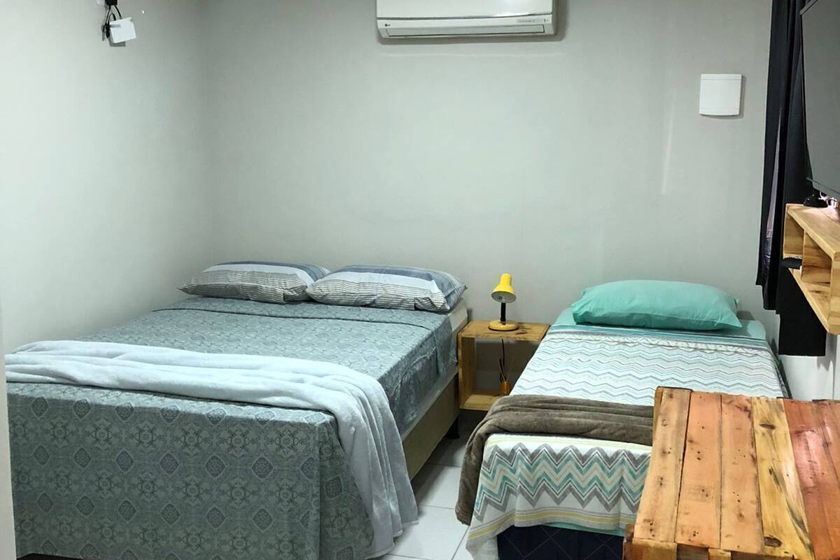 A bedroom with two beds, a wooden bench, a nightstand with a lamp, and an air conditioning unit.