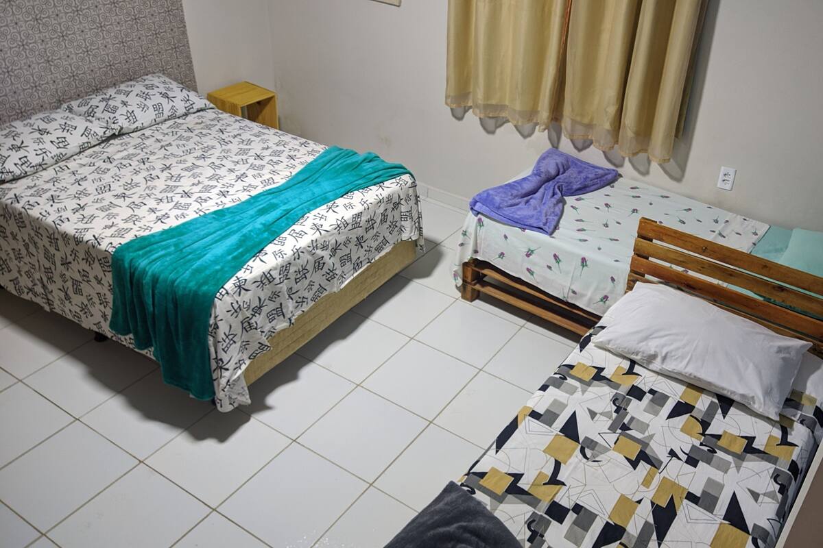 A room with two beds, one with a patterned bedspread and the other with a floral one. There is a small wooden stool and a window with curtains.