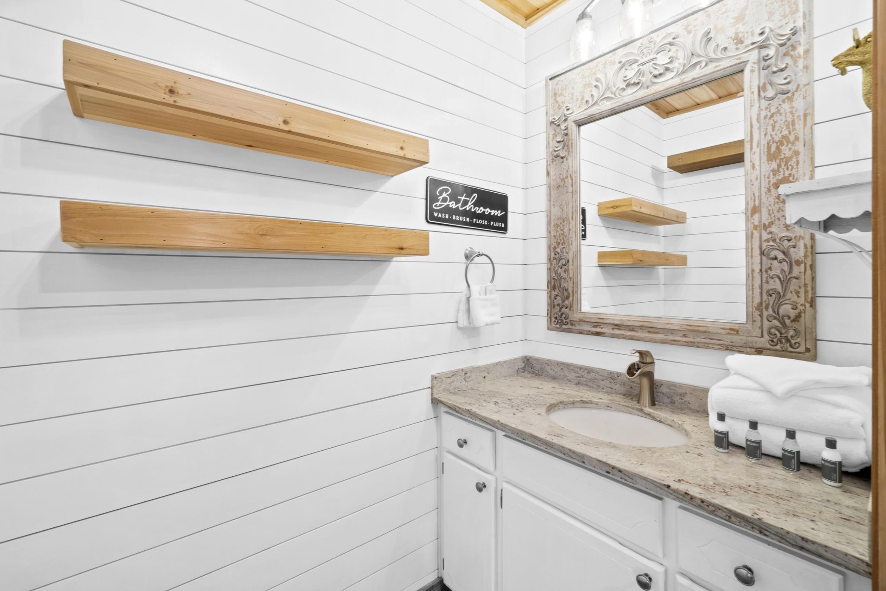 Une salle de bain avec un mur en bois peint en blanc, un lavabo surmonté d’un plan en marbre, un grand miroir avec un cadre décoratif et des étagères en bois au-dessus du lavabo.