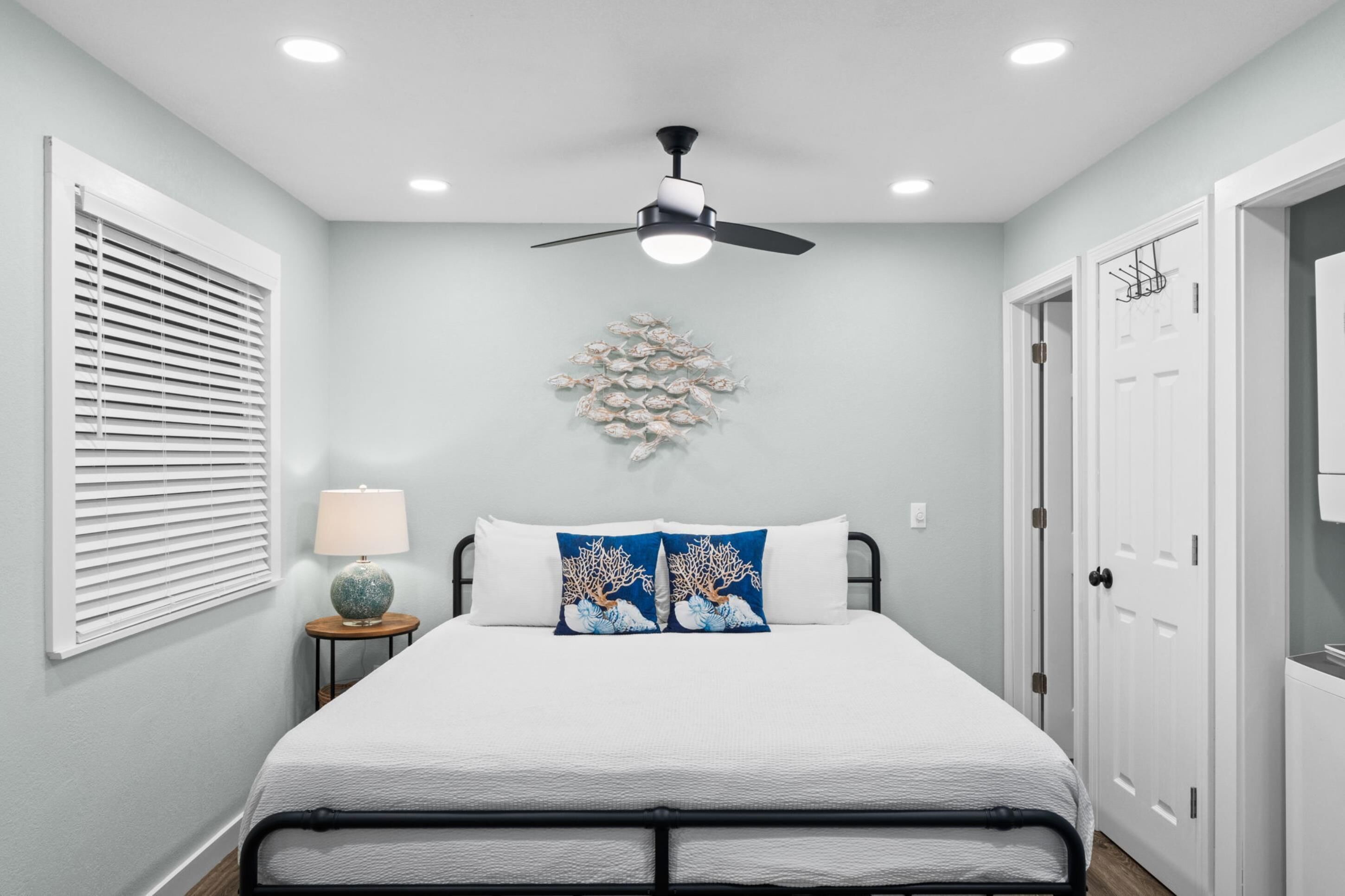 Un dormitorio con cama, ventilador de techo, luz empotrada en la pared, ventana con persianas y una pieza decorativa de arte mural con motivos de coral.