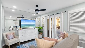 Uma sala de estar com televisão, ventiladores no teto, sofá e planta.