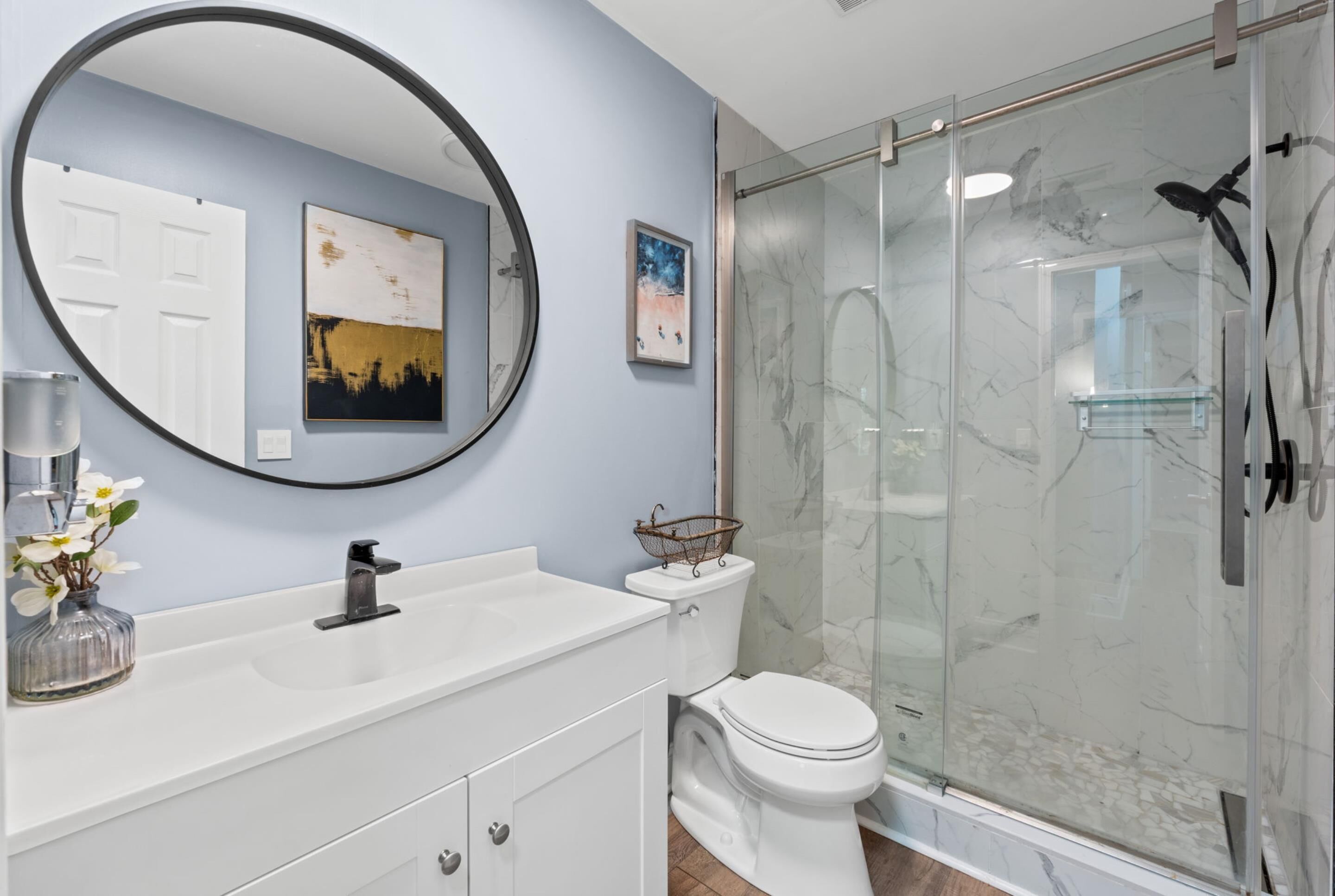A bathroom with a white vanity, a round mirror, a toilet, and a glass shower enclosure.