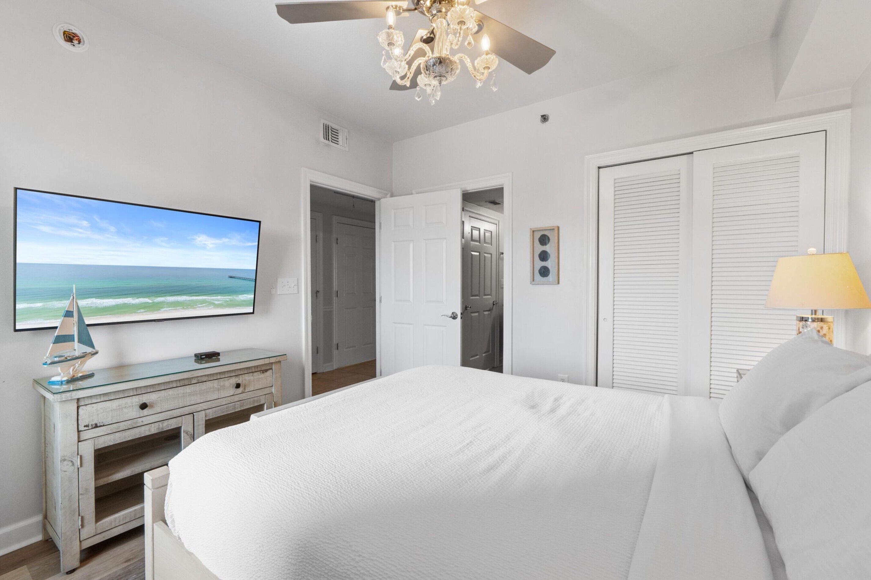 A bedroom with a bed, a television, a ceiling fan, a dresser, and a door leading to a bathroom.