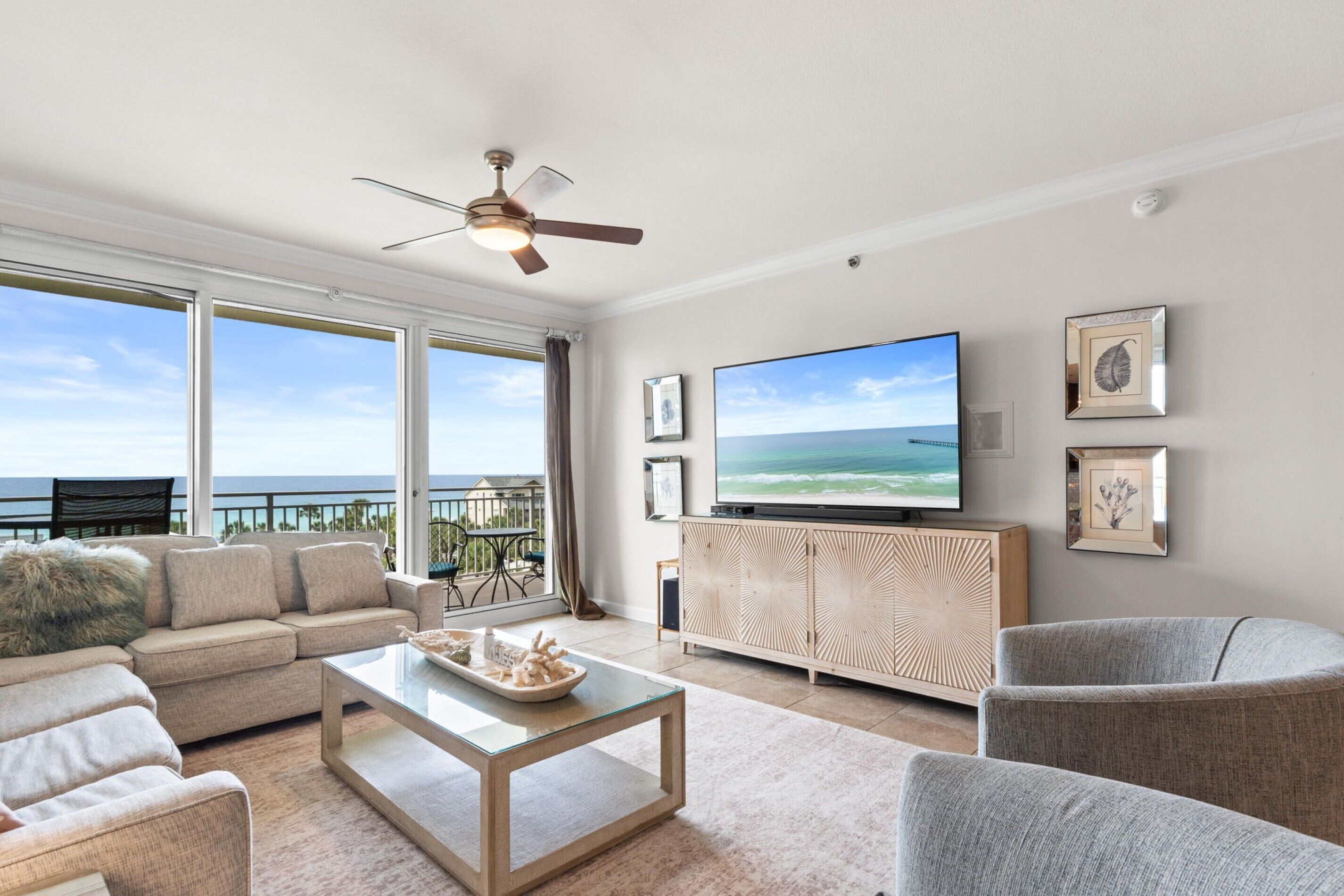Ein Wohnzimmer mit einem großen Fernseher, einem Deckenventilator, einer Couch, einem Couchtisch und Blick aufs Meer.