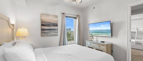 A hotel room with a bed, a television, a chandelier, a painting, a window with curtains, and a balcony.