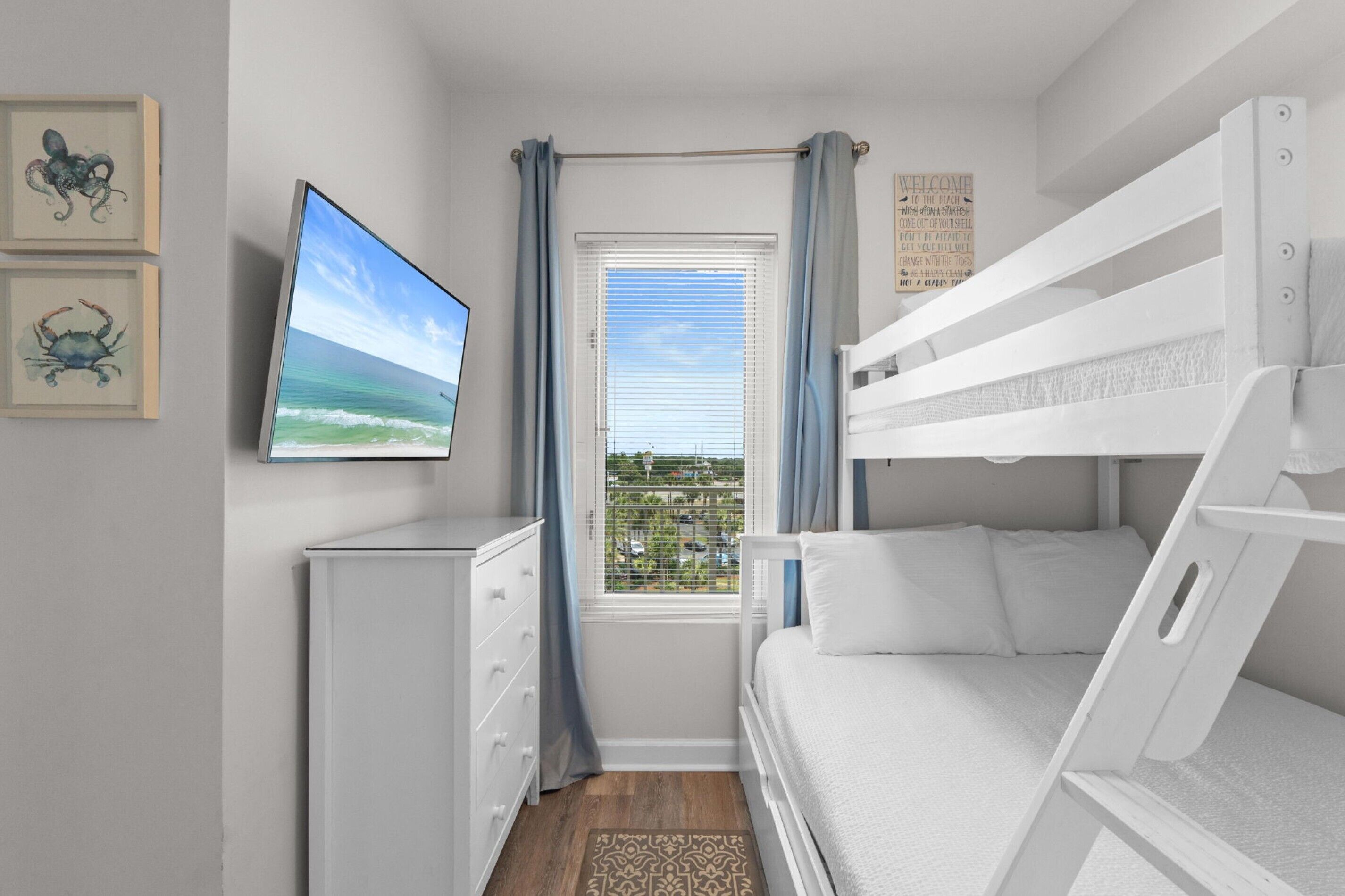 A bedroom with bunk beds, a TV, a dresser, a window with curtains, and framed artwork.