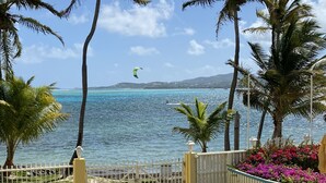A coastal view with a white fence, palm trees, and a clear sky. - Oceanfront, Remodeled 2BR- 2 King Size Beds / 2BA Condo at Ocean's View (Christiansted)
