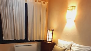 A neatly made bed with white and beige bedding, a wall-mounted light fixture, and a window with white curtains. - Oustaù des Carmes intra-muros (Avignon)