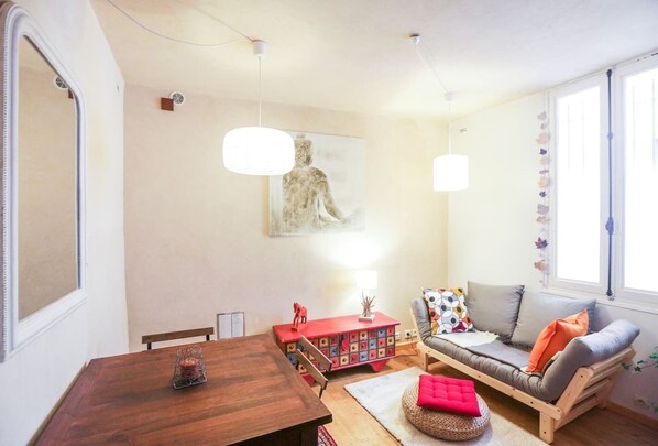 Een gezellige woonkamer met een houten tafel, een bank met kleurrijke kussens, een rode bijzettafel en een wandlamp.