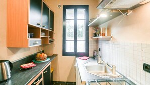A compact kitchen with a black-framed window, a stainless steel sink, and a countertop with a kettle and a basket of fruits. - Oustaù des Carmes intra-muros (Avignon)