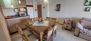Un coin repas avec une table en bois, des chaises et une nappe en dentelle. On aperçoit, au fond, un espace cuisine avec des placards et des appareils électroménagers.