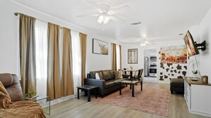 A modern living room with a sofa, coffee table, and a dining area with chairs. There is a ceiling fan, a TV mounted on the wall, and a kitchen visible in the background. - Low FEES! King Beds.  Free Parking, Wi-Fi, & Coffee (Westworth Village)