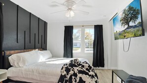 A bedroom with a bed, a ceiling fan, a window with curtains, a TV on the wall, and a bench. - Low FEES! King Beds.  Free Parking, Wi-Fi, & Coffee (Westworth Village)