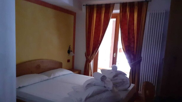 A hotel room with a wooden bed, a nightstand, a lamp, and a window with curtains. - Hotel dal Bracconiere (Dimaro Folgarida)