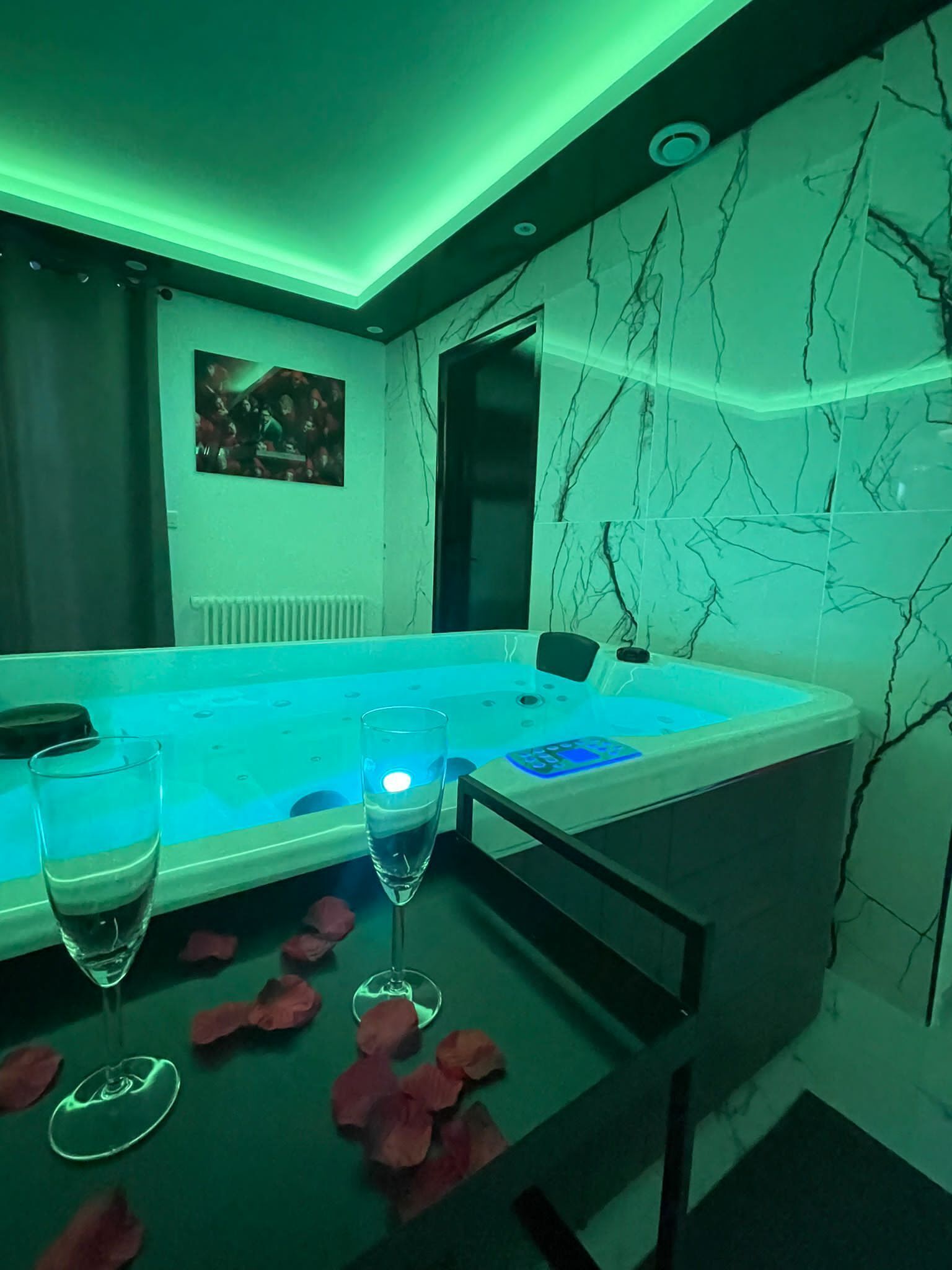 A modern spa room with a hot tub, two glasses, rose petals, and a marble wall.