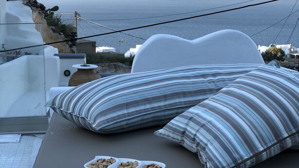 Un lit avec des coussins à rayures et un bol de noix sur un balcon donnant sur une falaise et la mer.