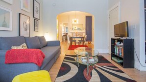 A living room with a sofa, a coffee table, a bookshelf, and a TV. - Elmwood Village 2BR Apt •Sleeps 8• Walk to Dining (Buffalo)