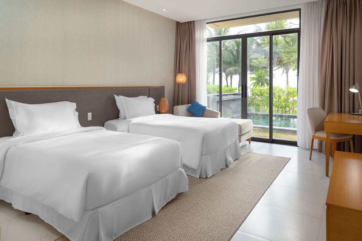 A hotel room with two beds, a desk, and a view of greenery outside.