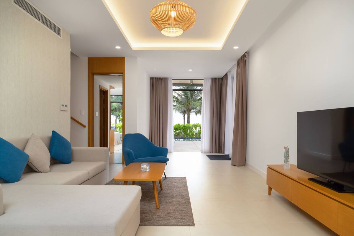 A modern living room with a sofa, blue armchair, coffee table, TV, and a view of palm trees through large windows.