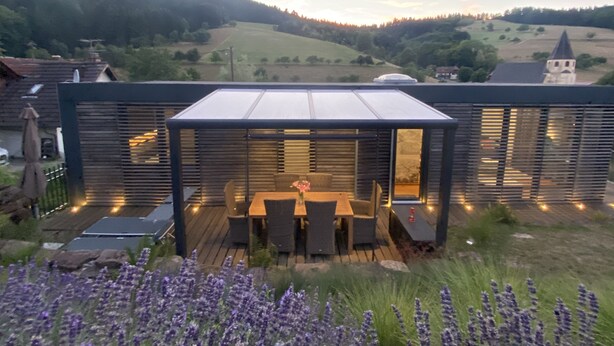 Une terrasse extérieure moderne, dotée d’une véranda en verre, d’une terrasse en bois et d’une table avec des chaises.